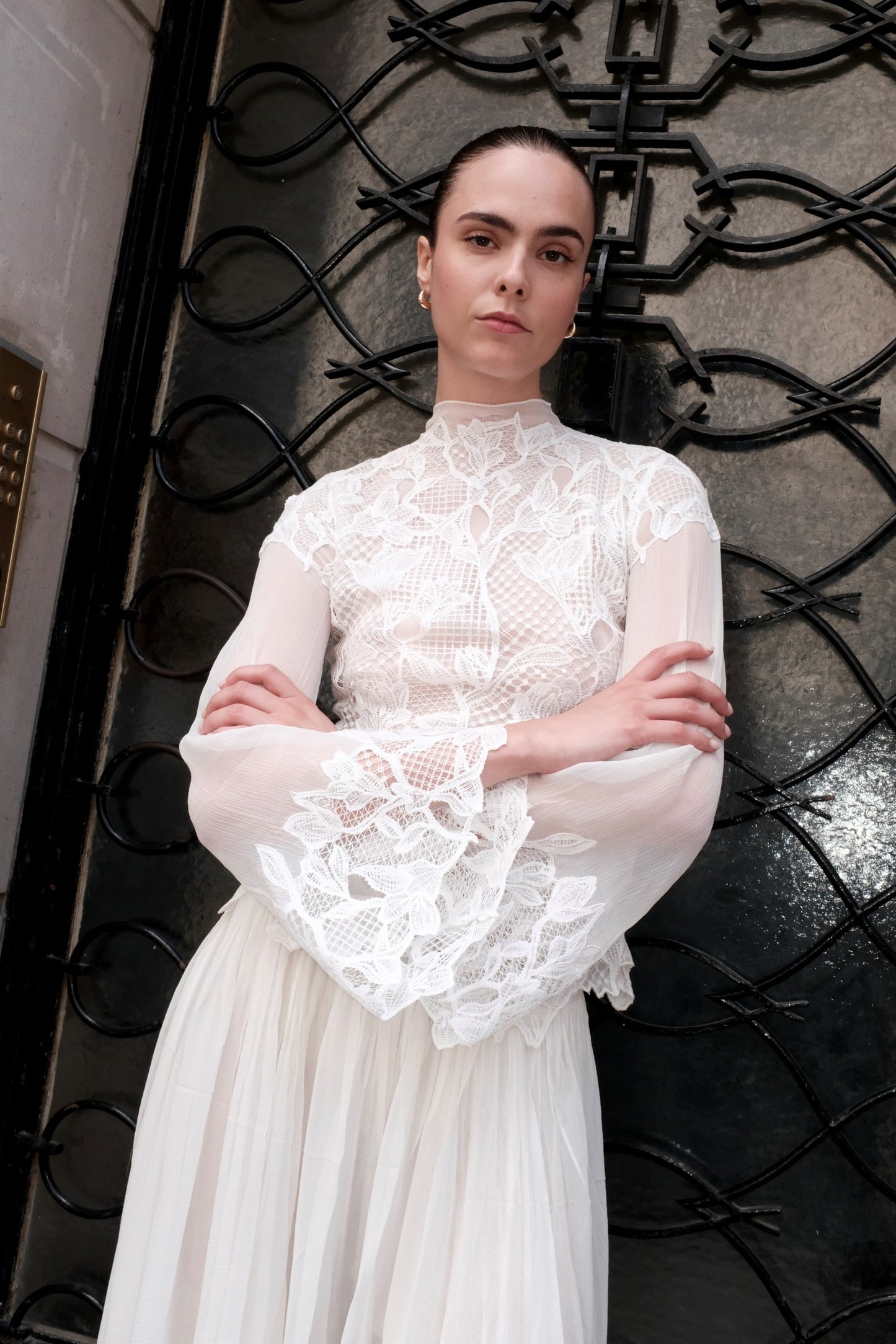 A woman wearing a white lace and mesh long-sleeve dress with a high neck and a flowing skirt, standing in front of a black wrought-iron gate.