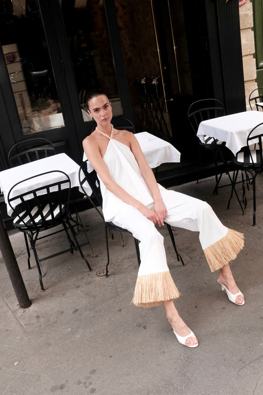A woman sits on a black metal cafe chair outside a restaurant, wearing a white halter-neck jumpsuit with fringed flared hems and white heeled sandals.