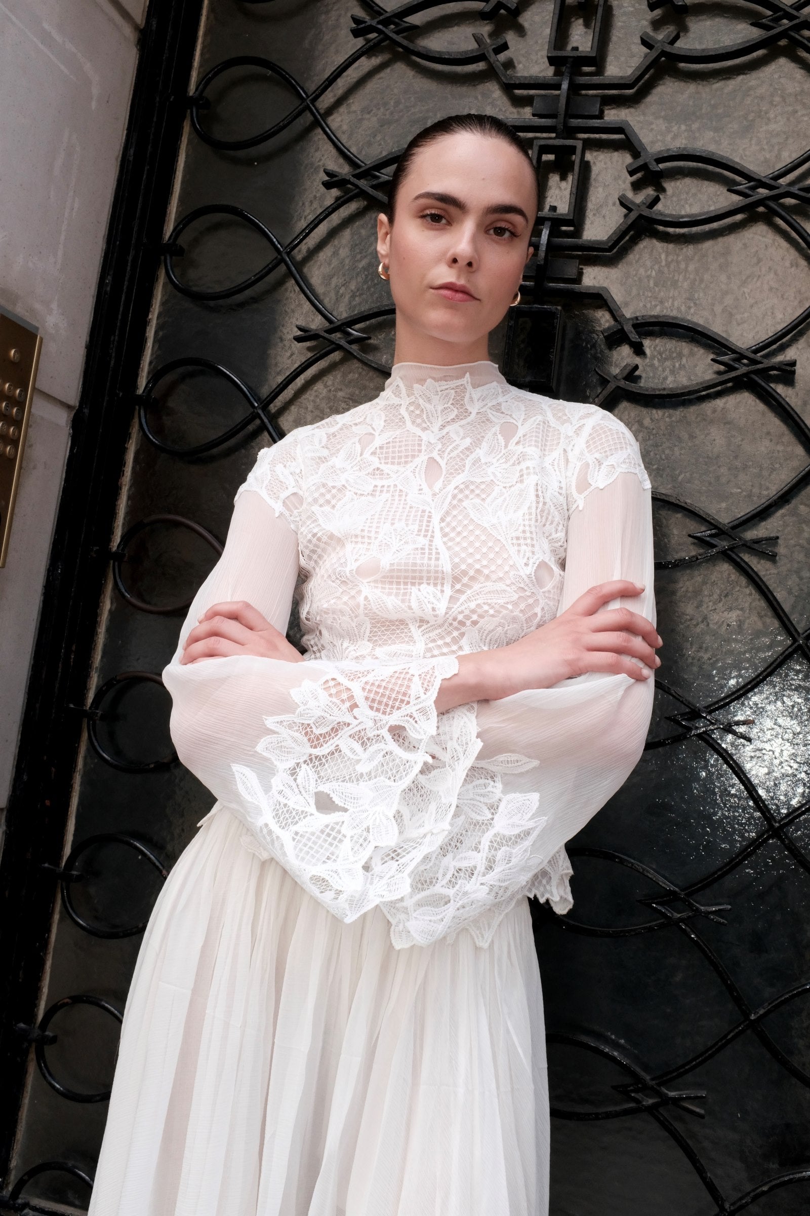 A woman wearing a white lace and mesh long-sleeve dress with a high neck and a flowing skirt, standing in front of a black wrought-iron gate.