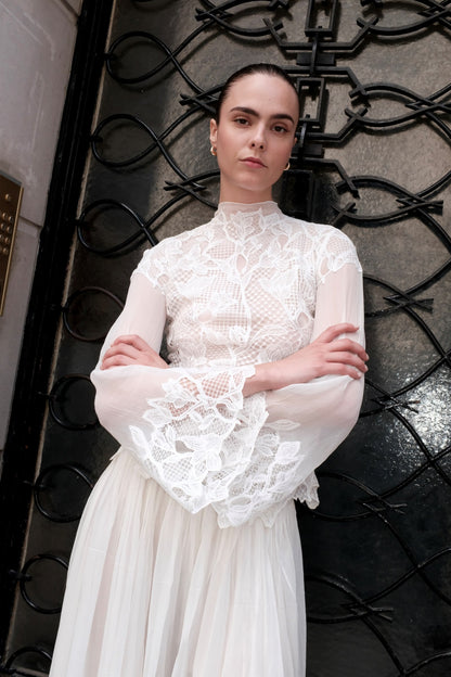 A woman wearing a white lace and mesh long-sleeve dress with a high neck and a flowing skirt, standing in front of a black wrought-iron gate.
