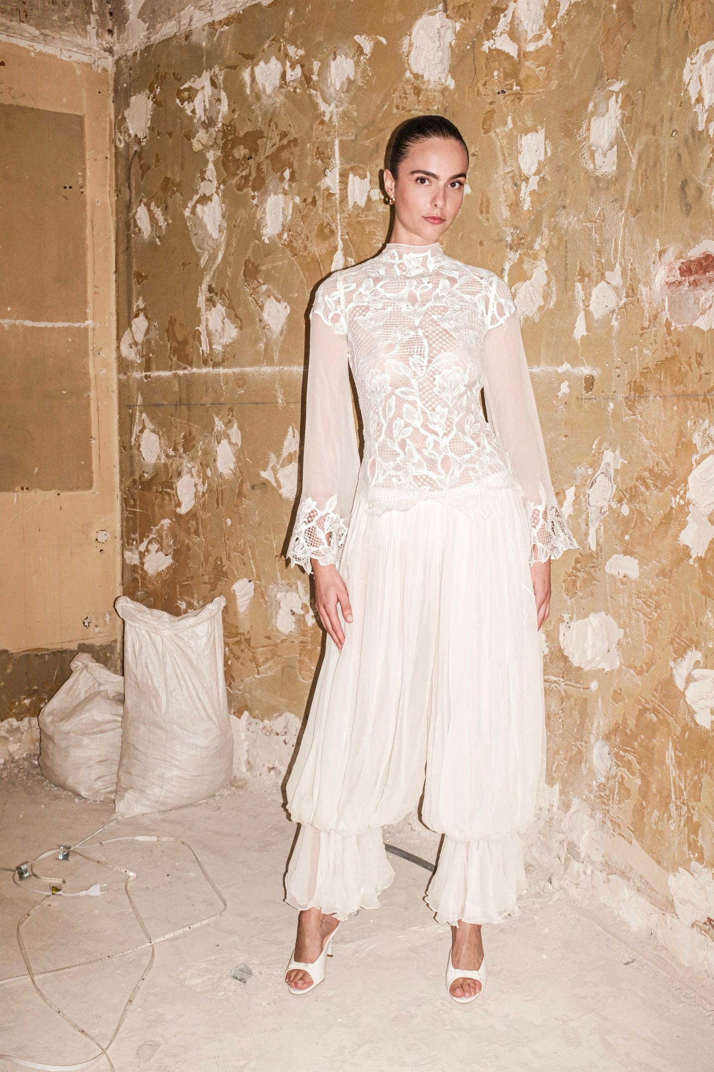 A model stands in a distressed, industrial-style room with peeling beige and brown walls, wearing a cream-colored two-piece outfit consisting of a high-neck lace long-sleeve top and matching pleated wide-leg pants.