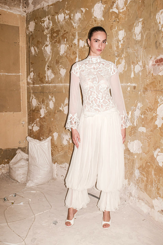 A model stands in a distressed, industrial-style room with peeling beige and brown walls, wearing a cream-colored two-piece outfit consisting of a high-neck lace long-sleeve top and matching pleated wide-leg pants.